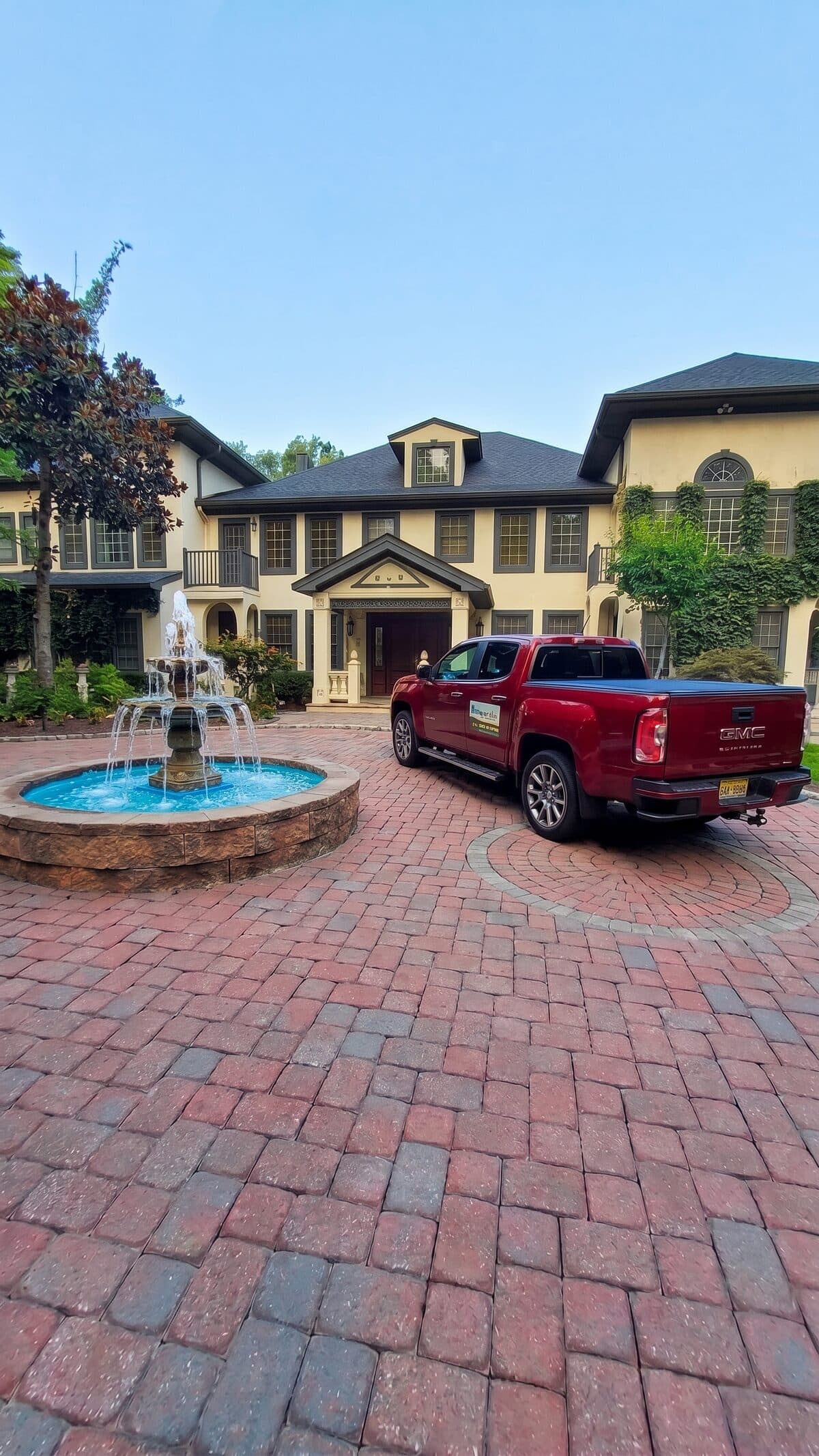 The red HomeCheck truck parked on a paver driveway beside a fountain, in front of a large New Jersey home